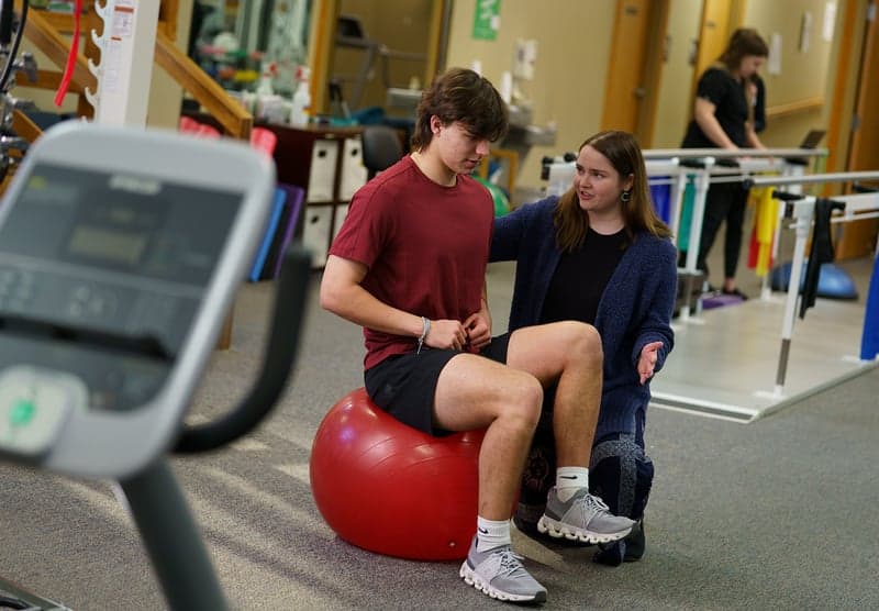 A young male sits on an exercise ball with a physical therapist