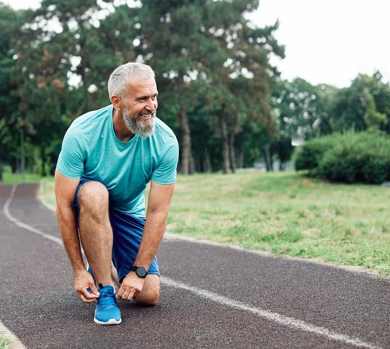 A man with grey hair kneels on a trail to tie his shoe