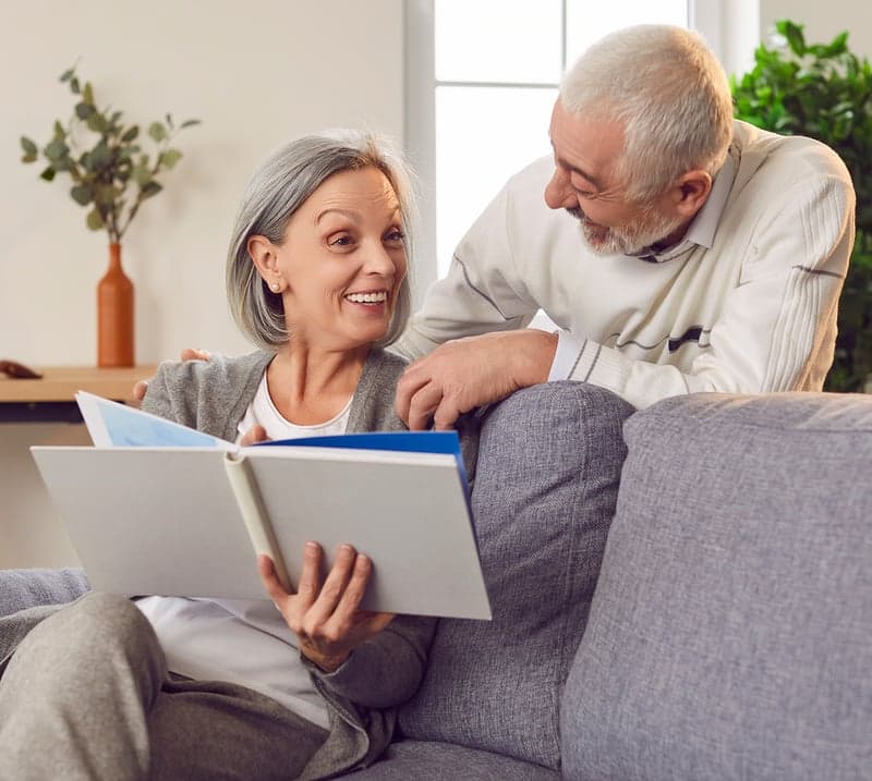 An older woman sits on a couch showing a book to a mature man standing behind her