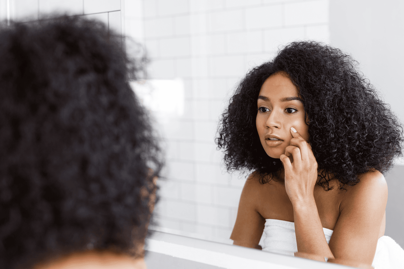 A young woman looking at mirror and examines her skin, touching face with finger